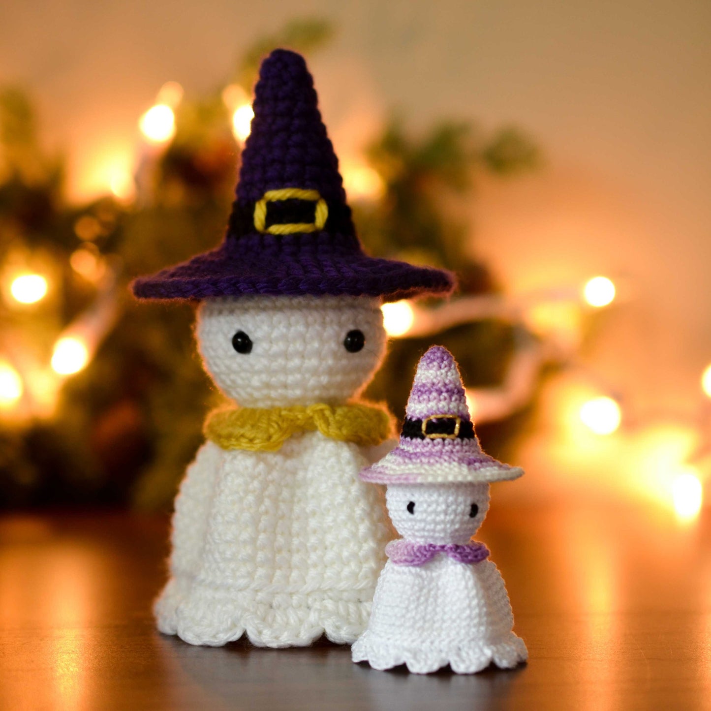 Two crocheted ghost figures wearing purple and white witch hats. The larger ghost has a yellow collar and the smaller ghost has a purple collar. The ghosts are displayed against Halloween-themed background.