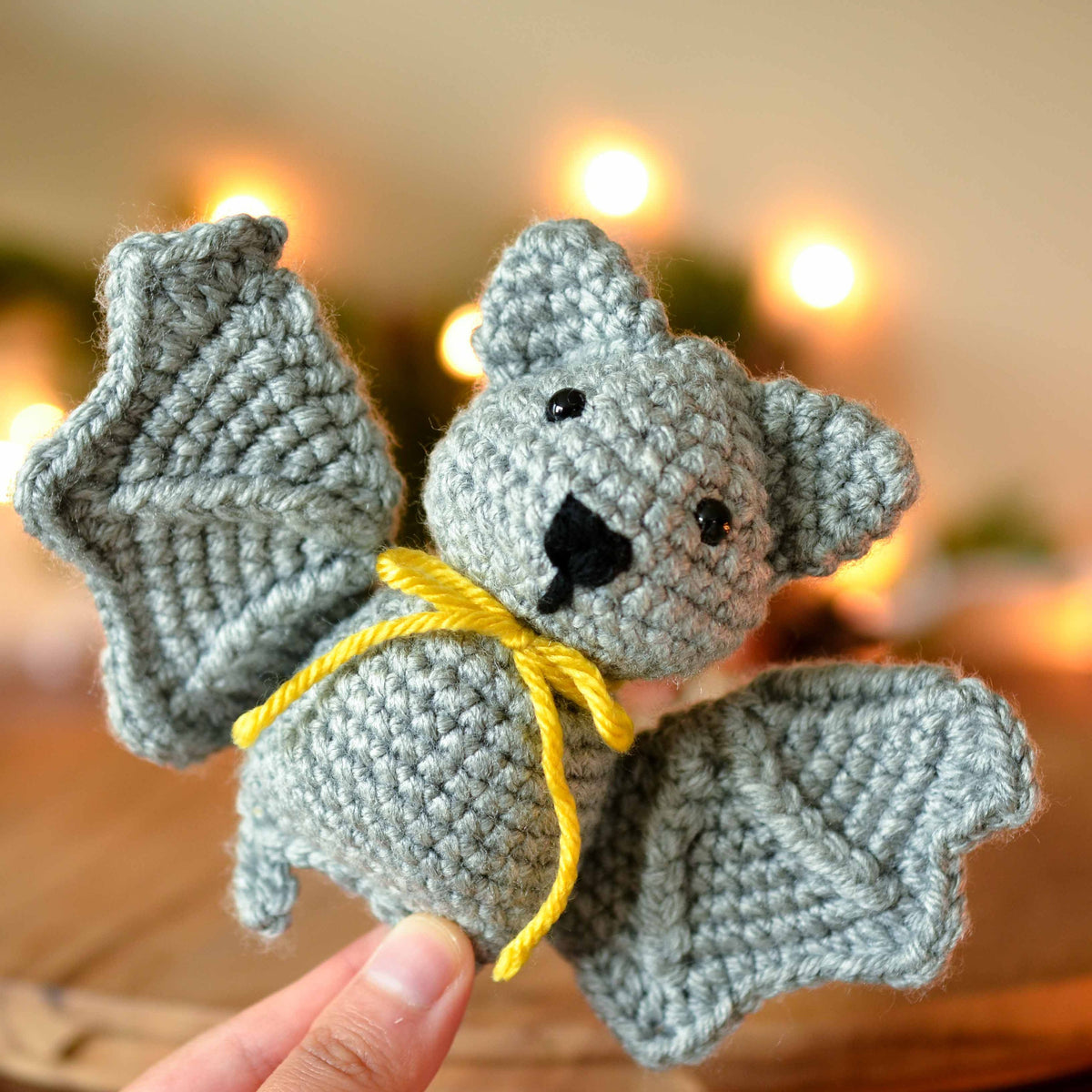 A gray crocheted bat with yellow yarn tied around its neck. The bat has black eyes and a black nose.