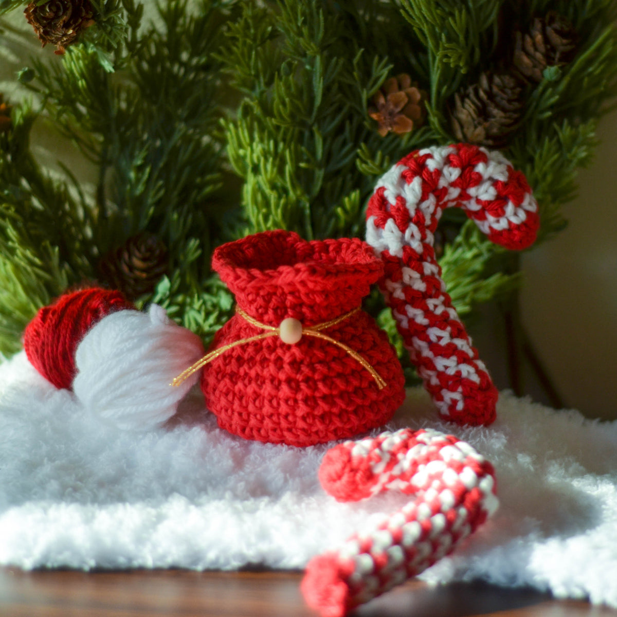 A red crocheted holiday pouch with a gold ribbon and a wooden bead. Next to the pouch is a white and red crocheted candy cane. There is another white and red crocheted candy cane in the foreground.