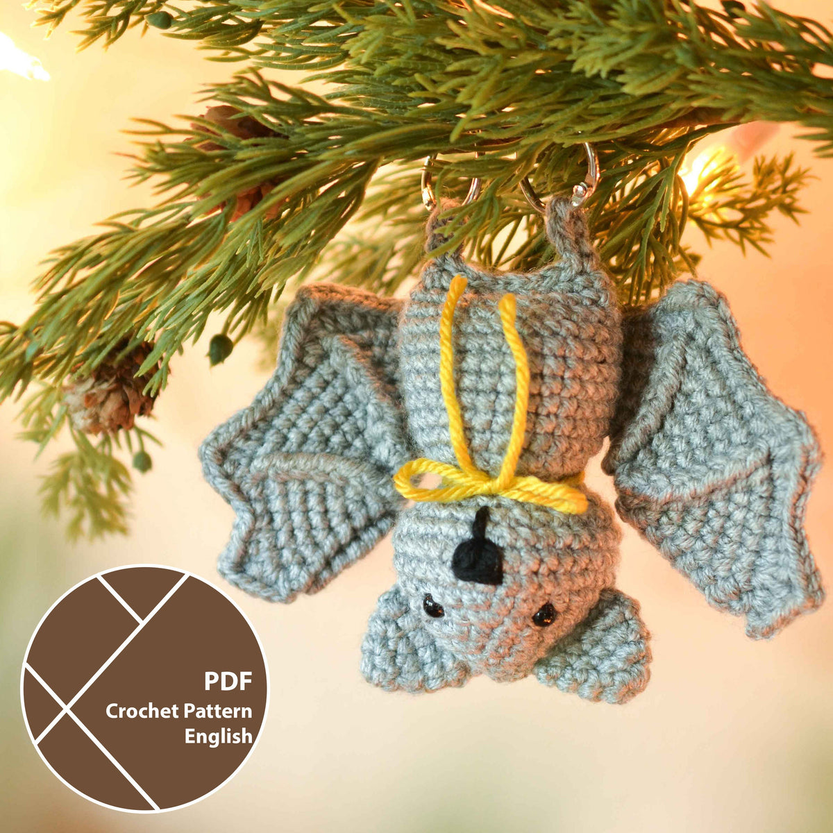 A gray crocheted bat ornament with yellow yarn tied around its neck. The bat is hanging from a tree branch.