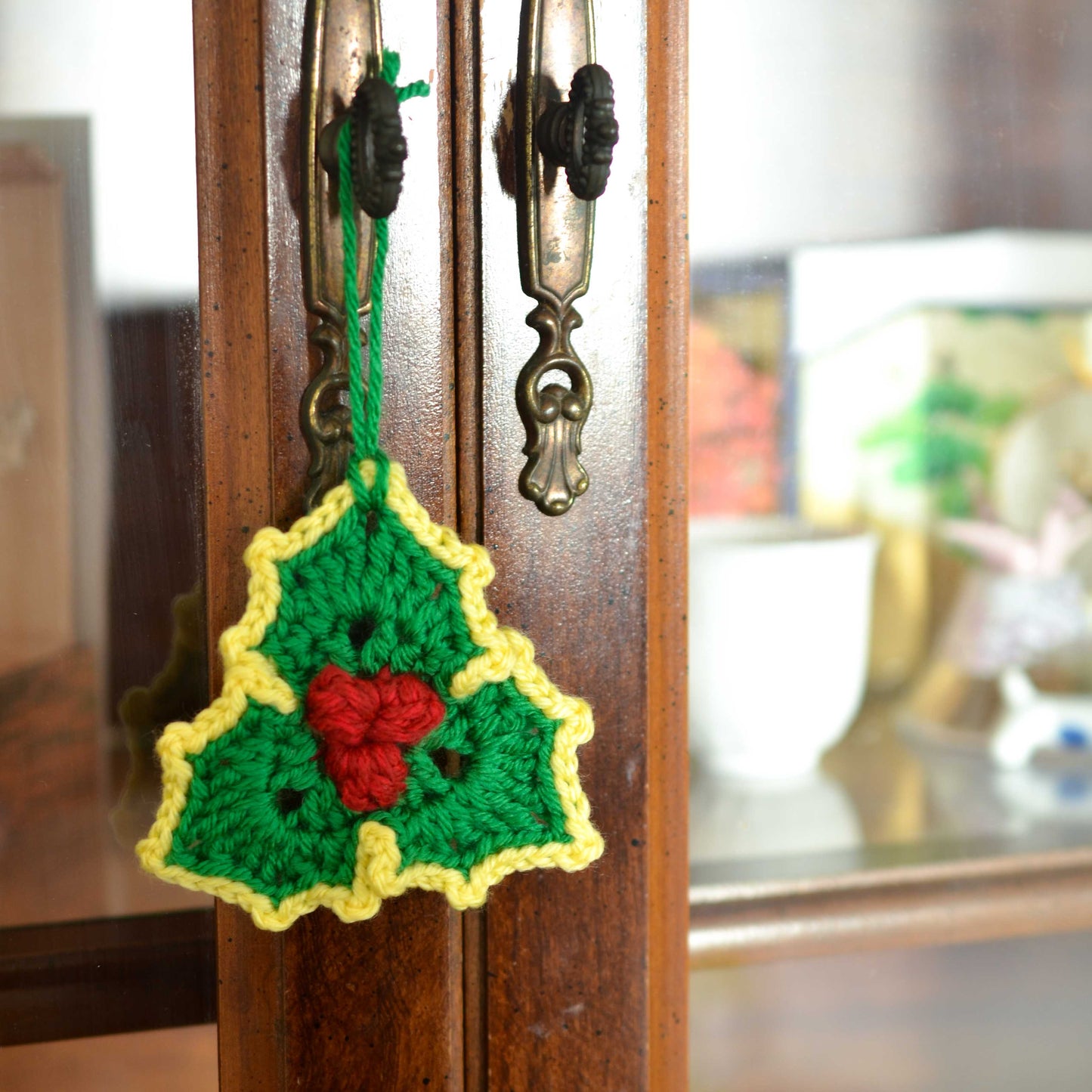 A crocheted holly leaf Christmas ornament. It is hanging from a dark brown wooden cabinet. It is a festive decoration perfect for the holiday season. The crocheted holly leaf ornament is a unique and charming addition to any Christmas tree or home decor.