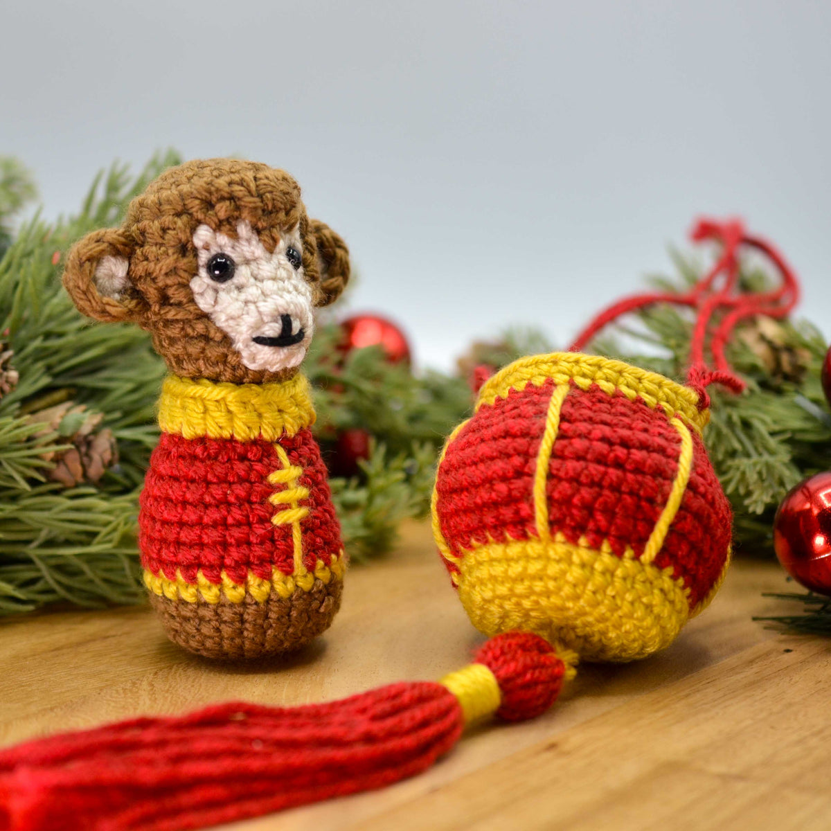 A crocheted monkey, wearing Lunar-New-Year clothing, is sitting on a wooden surface, facing right. A crocheted red lantern is next to the monkey.