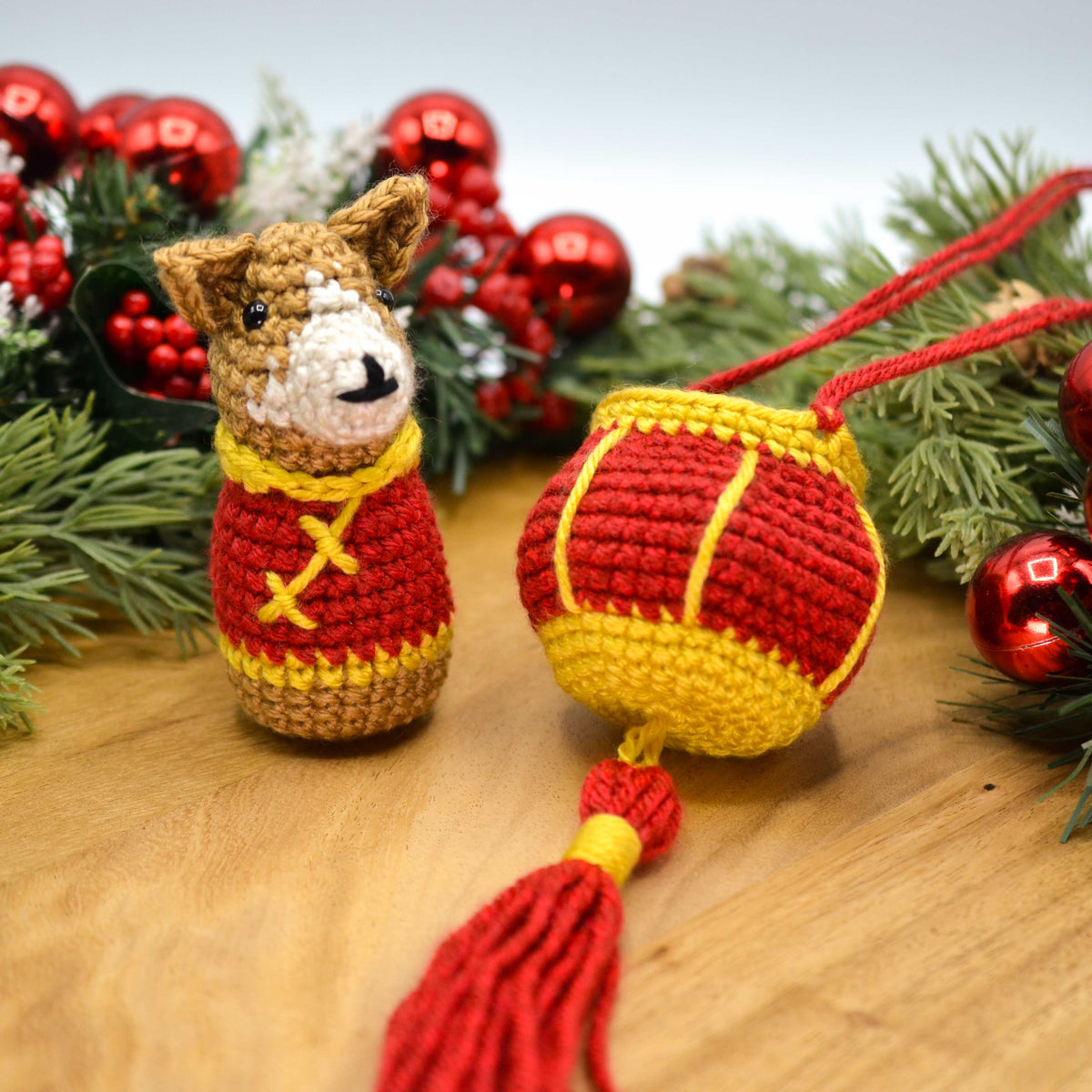 A crocheted dog, wearing Lunar-New-Year clothing, is sitting on a wooden surface. A crocheted red lantern is next to the dog.