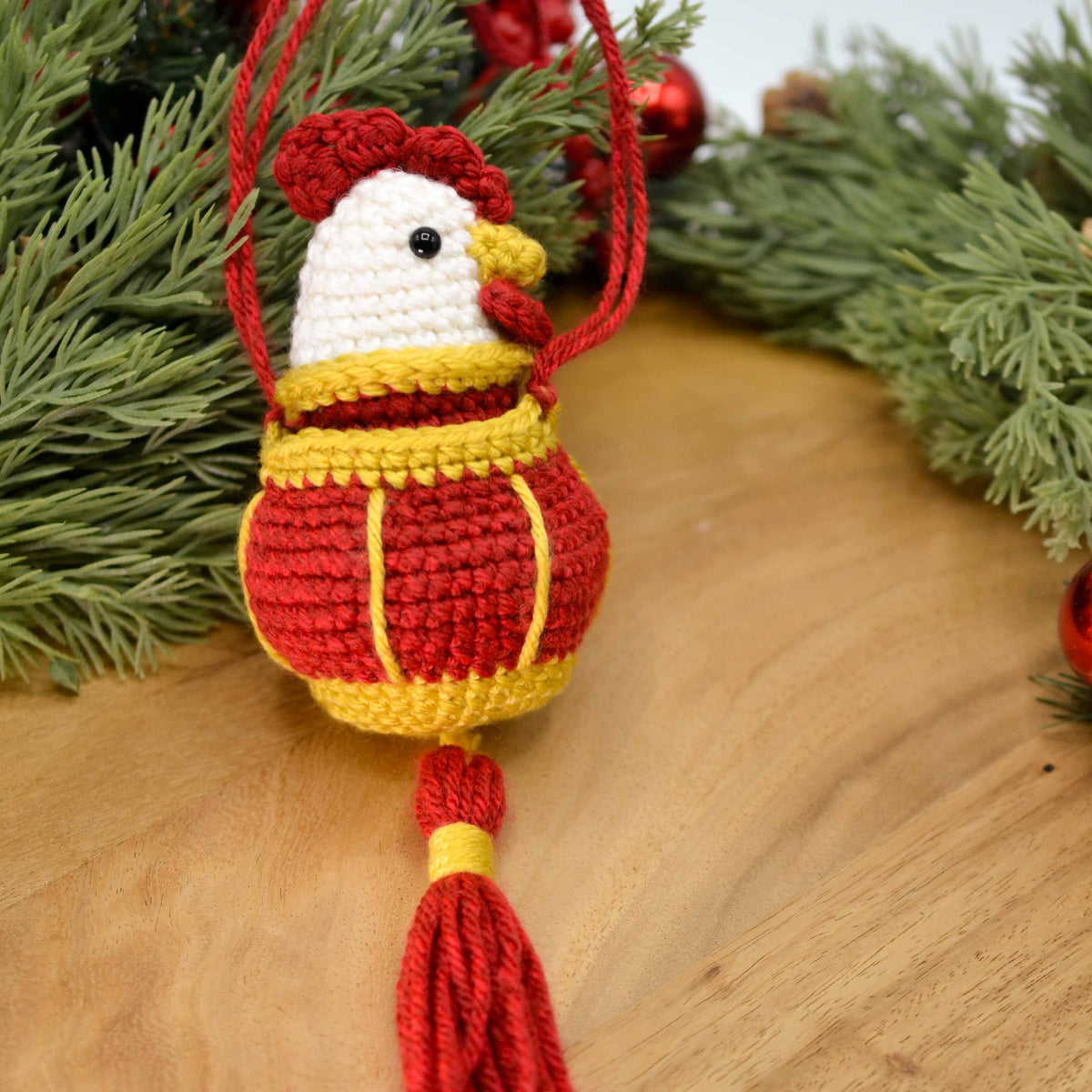 A crocheted rooster is placed in a crocheted red lantern. The set is displayed on a wooden surface against some green branches.