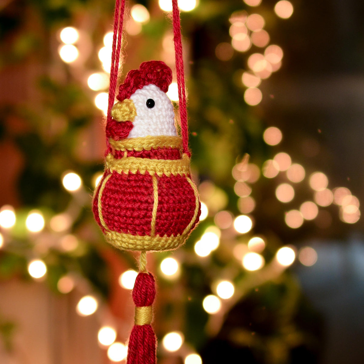 Crocheted rooster ornament against a blurred Christmas tree background.