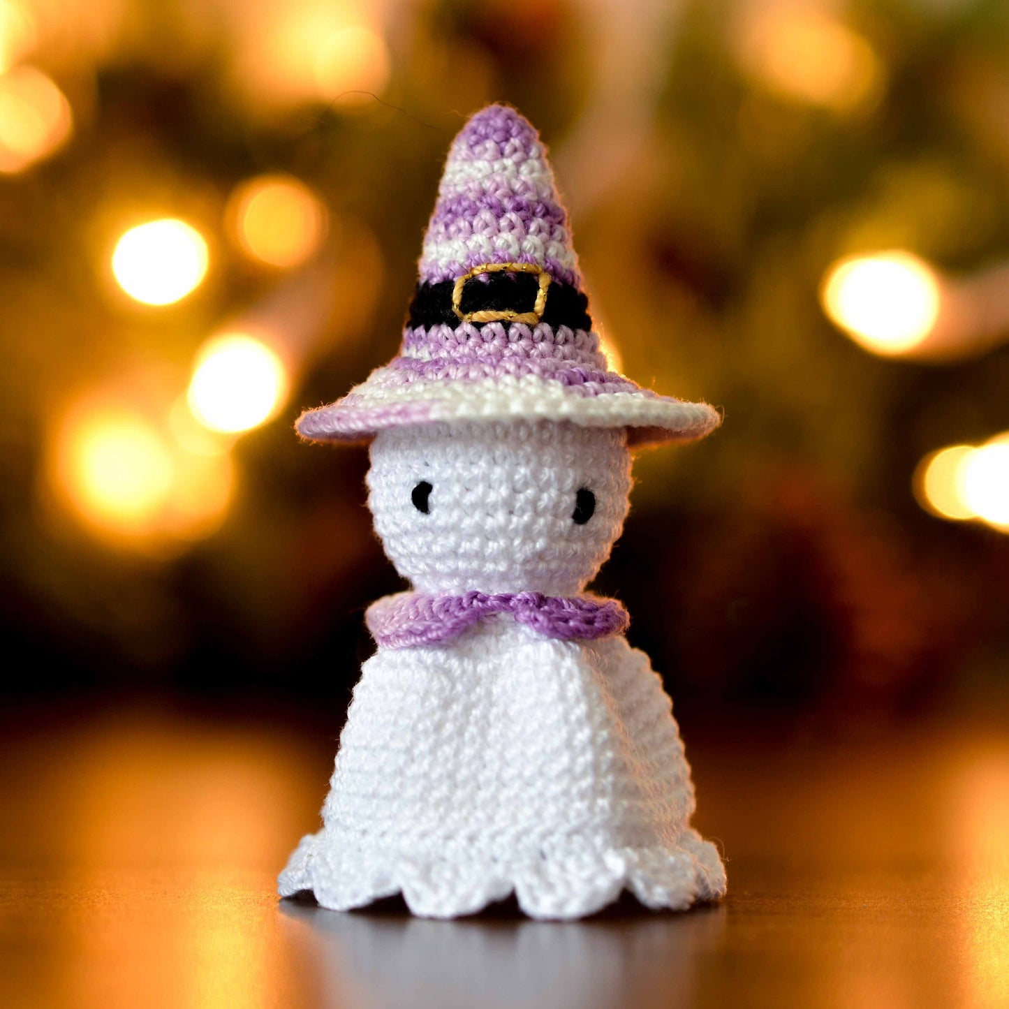 A crocheted ghost wearing a purple and white striped witch hat. The ghost is white and has a purple collar around its neck.