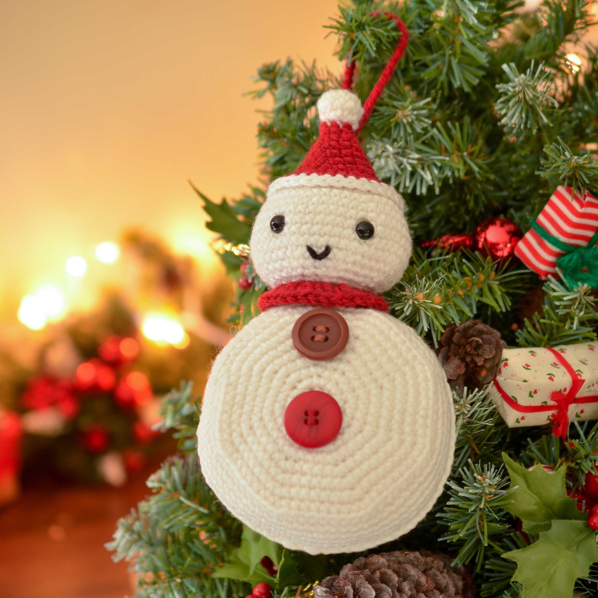 A white crocheted snowman ornament with a red hat, red scarf, and buttons. The snowman is hanging on a green Christmas tree with red and green ornaments.