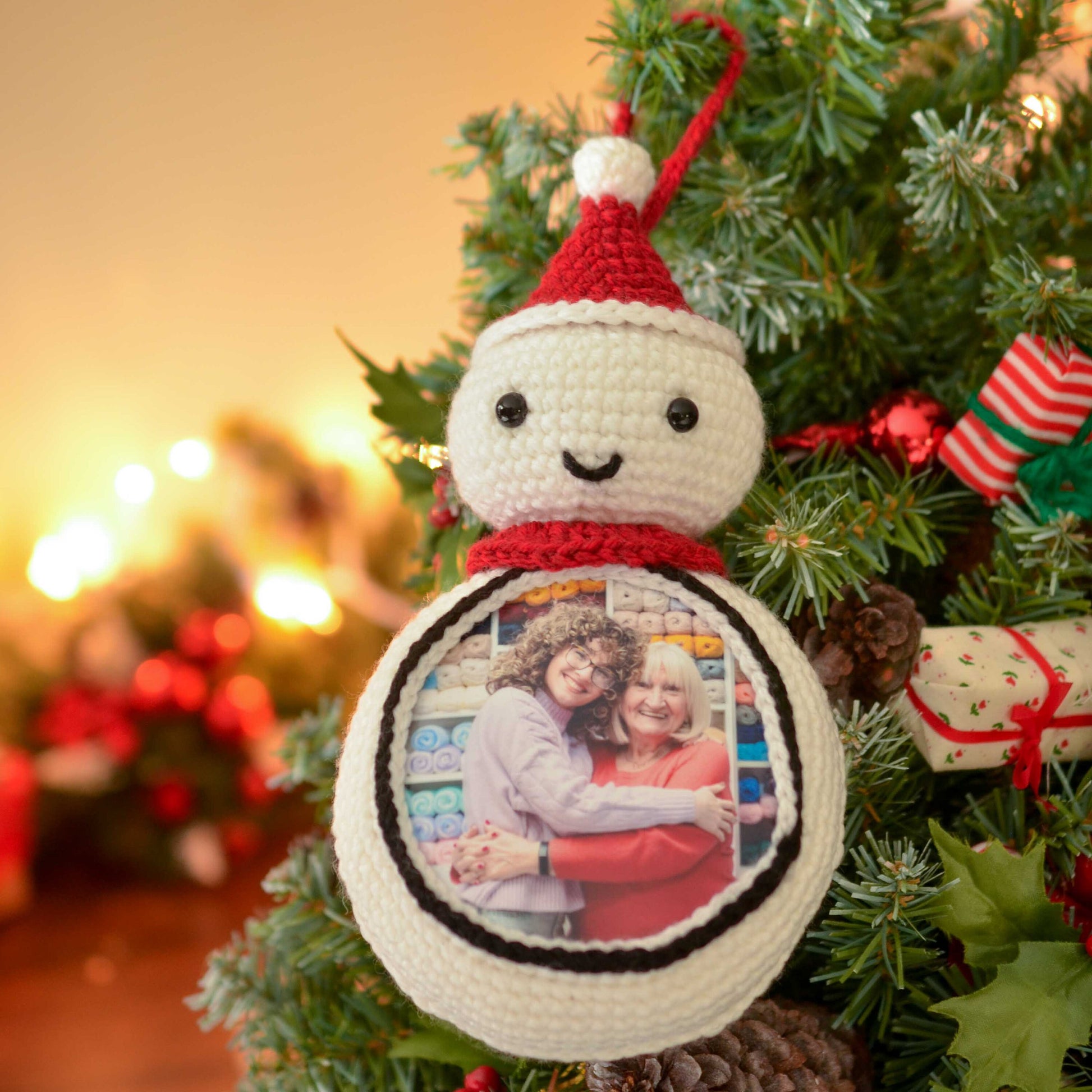 A white crocheted snowman ornament with a red Santa hat and a photo frame in the front. The photo frame contains a picture of two people smiling and hugging. The ornament is hanging on a green Christmas tree.