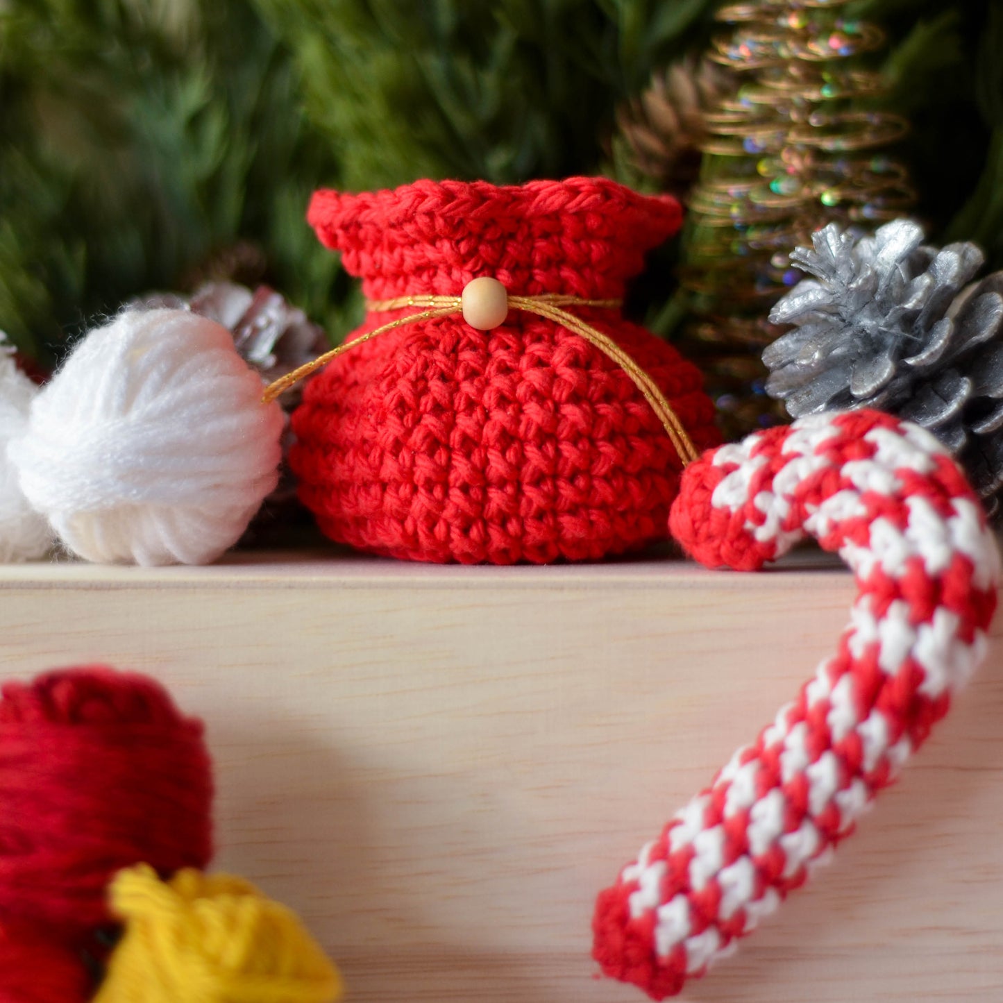 A red crocheted holiday pouch with a gold drawstring and a wooden bead. A white and red crocheted candy cane is next to the pouch.