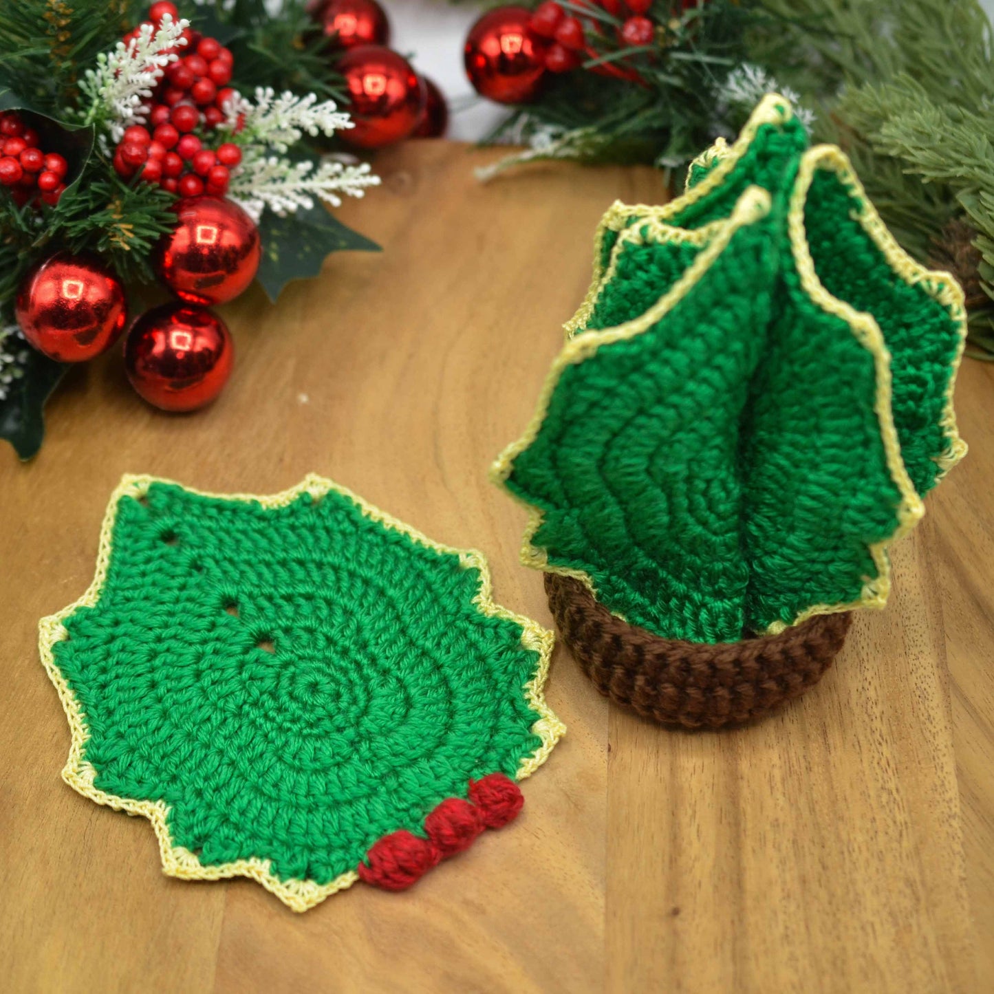 Crocheted Holly Christmas Tree Coaster Set. One holly leaf coaster is unfolded and displayed on a wooden surface. The rest of the coasters are store in a darn brown holder. The stored coasters in a holder looks like a Christmas tree. 