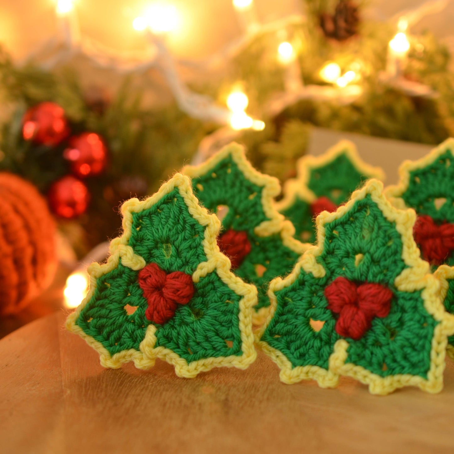 Several crocheted holly leaf ornaments. Each ornament is green with yellow edges and features red crocheted berries in the center. The ornaments are arranged on a wooden surface against a blurred background of Christmas lights and greenery.