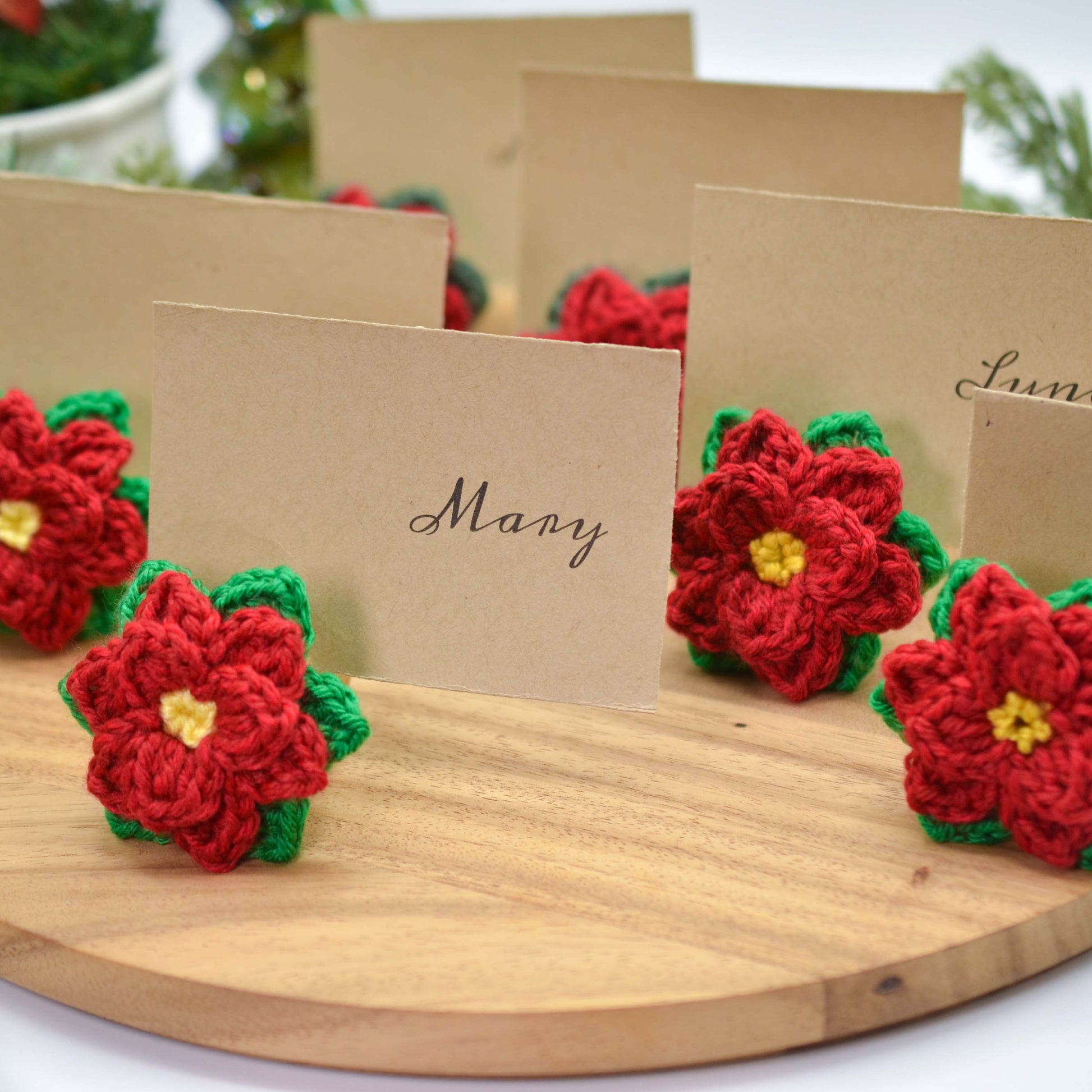 Crochet pattern for poinsettia place card holders. Image shows several red and green crocheted poinsettias attached to place cards with names written on them.  The place cards are brown and the poinsettias are a festive Christmas decoration.