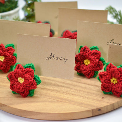 Crochet pattern for poinsettia place card holders. Image shows several red and green crocheted poinsettias attached to place cards with names written on them.  The place cards are brown and the poinsettias are a festive Christmas decoration.