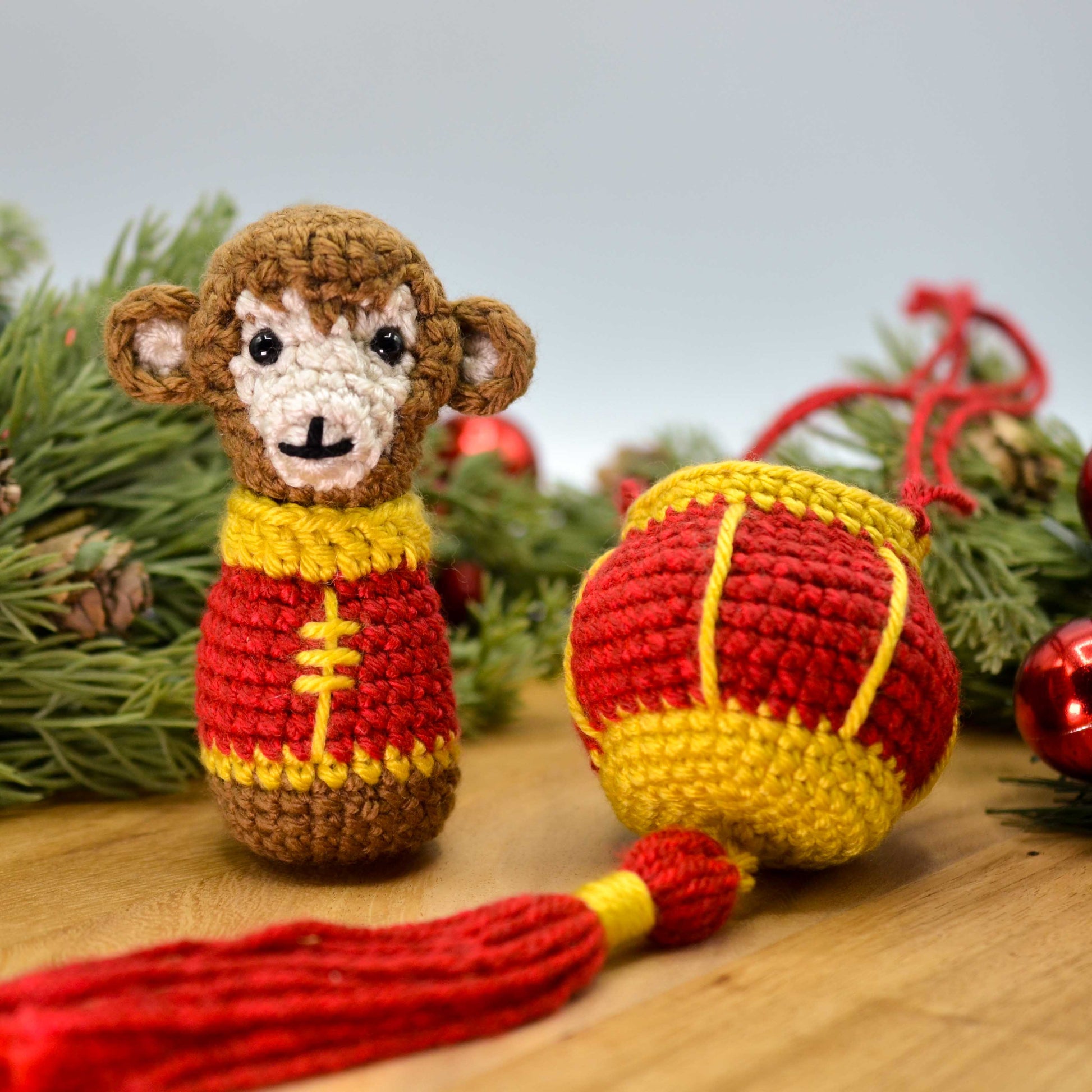 A crocheted monkey, wearing Lunar-New-Year clothing, is sitting on a wooden surface, facing front. A crocheted red lantern is next to the monkey.
