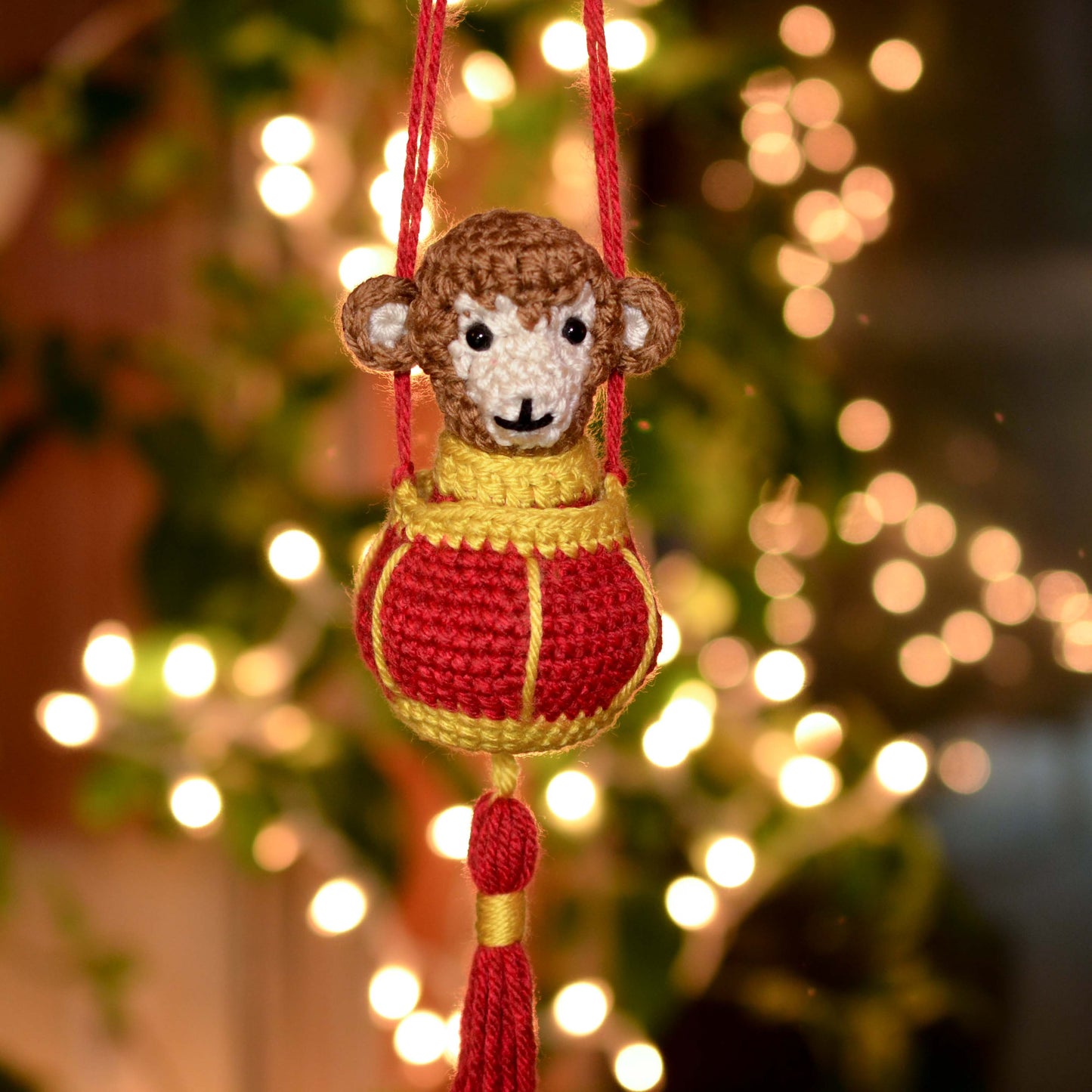 A crocheted monkey is sitting in a crocheted Lunar New Year red lantern. The set is hanging in front of a background with holiday lighting. 
