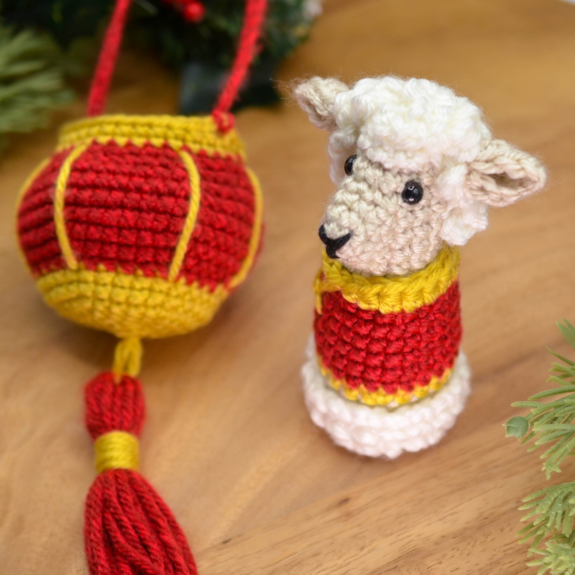 A crocheted sheep is in Lunar-New-Year style clothing, and a crocheted red lantern is placed next to the sheep. The hanging ornament set is placed on a wooden surface.