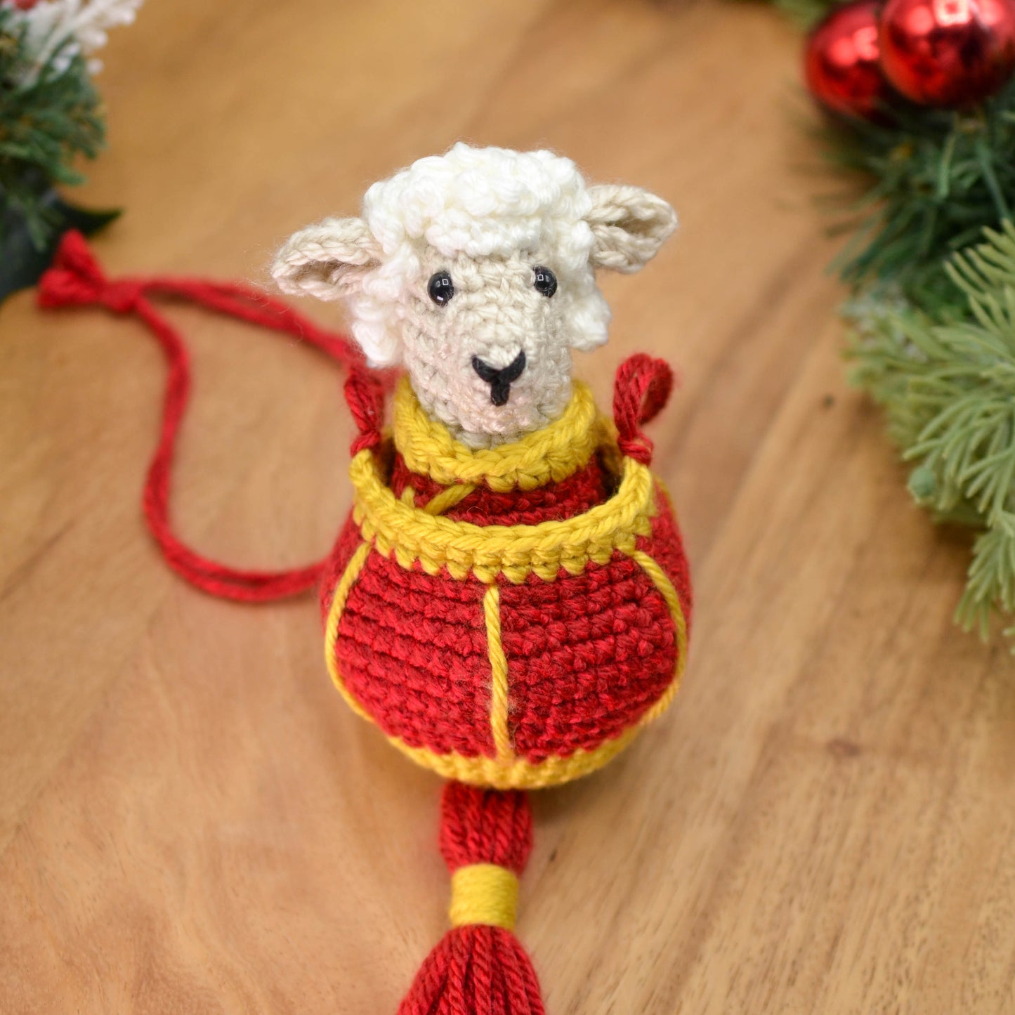 A crocheted sheep wearing Lunar-New-Year style clothing, and it's placed in a crocheted red lantern. The hanging ornament set is placed on a wooden surface.