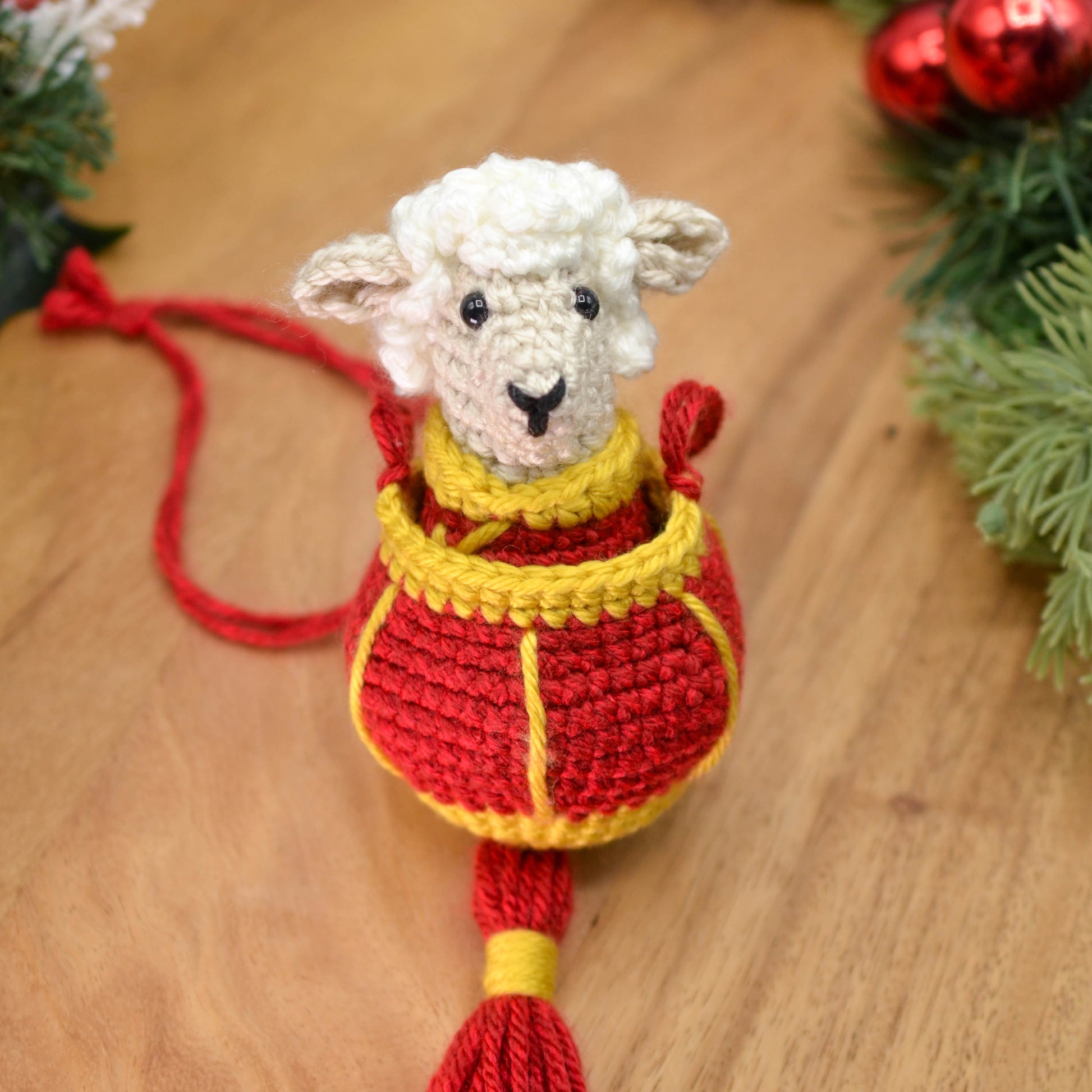 A crocheted sheep wearing Lunar-New-Year style clothing, and it's placed in a crocheted red lantern. The hanging ornament set is placed on a wooden surface.