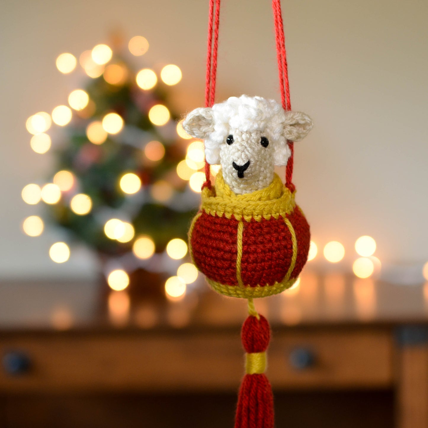 A crocheted sheep sitting in a red lantern basket. The set is a hanging ornament that is currently hanging in front of a festive background.