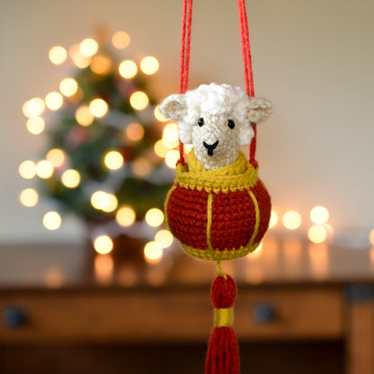 A crocheted sheep sitting in a red lantern basket. The set is a hanging ornament that is currently hanging in front of a festive background.