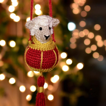 A crocheted sheep wearing Lunar-New-Year style clothing, and it's placed in a crocheted red lantern. The hanging ornament set is displayed against a dark background with festive lighting.