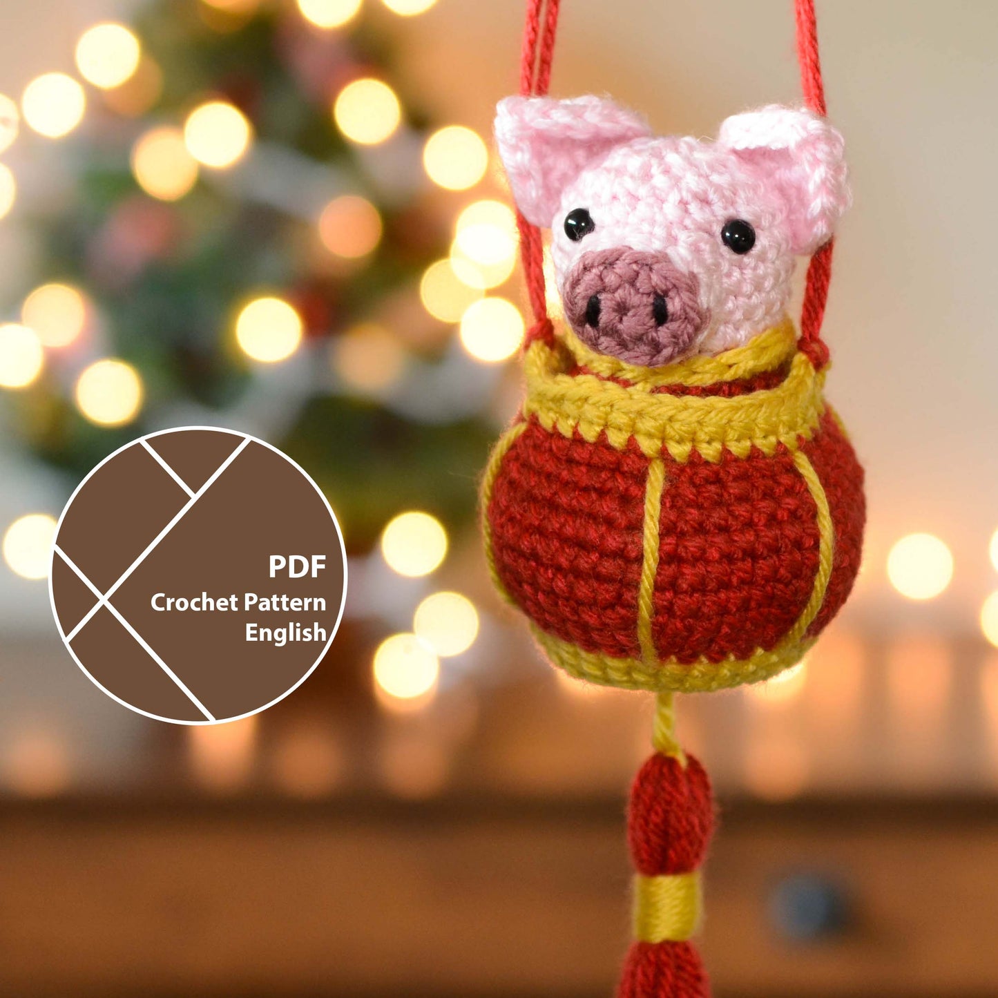 Crochet pattern for a pig ornament and a red lantern in front of a blurred festive background. A label indicates that this listing is a crochet pattern PDF, written in English. 
