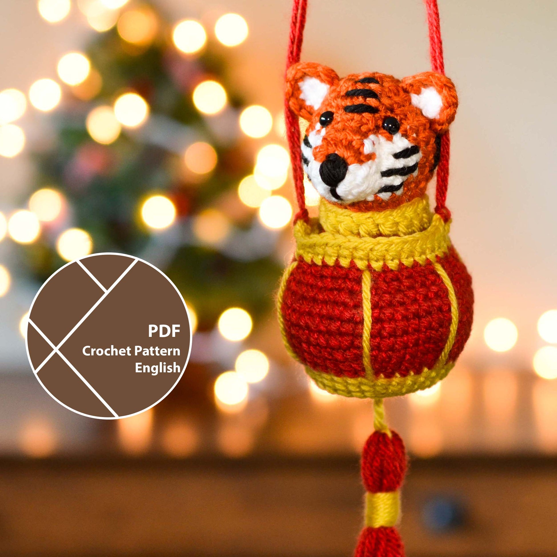 crocheted tiger ornament with red lantern hanging in front of a festive background; a label indicates that this listing is a crochet pattern PDF written in English. 