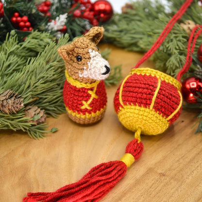 A crocheted dog, wearing Lunar-New-Year clothing and facing right, is sitting on a wooden surface. A crocheted red lantern is next to the dog. 
