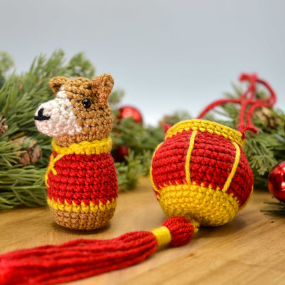 A crocheted dog, wearing Lunar-New-Year clothing and facing left, is sitting on a wooden surface. A crocheted red lantern is next to the dog.