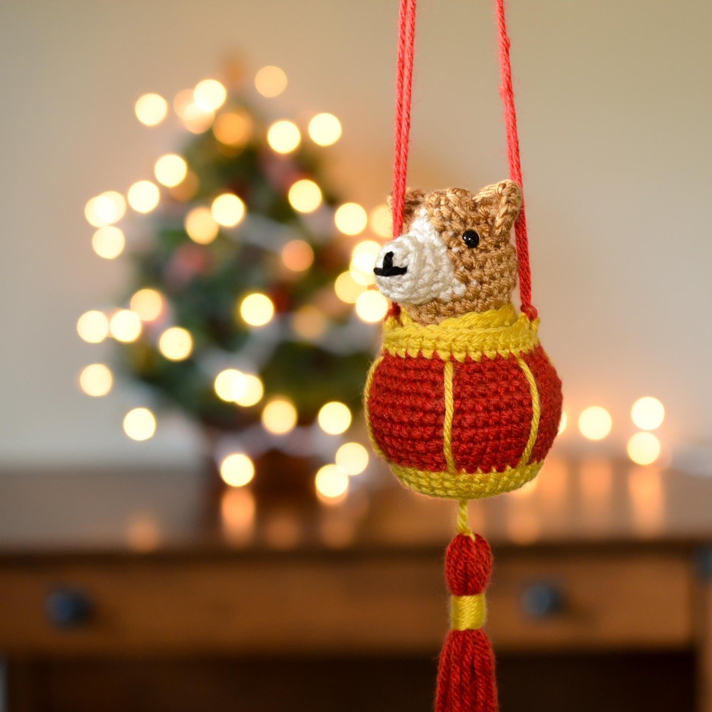 A crocheted dog and red lantern ornament is hanging in front of a festive background.