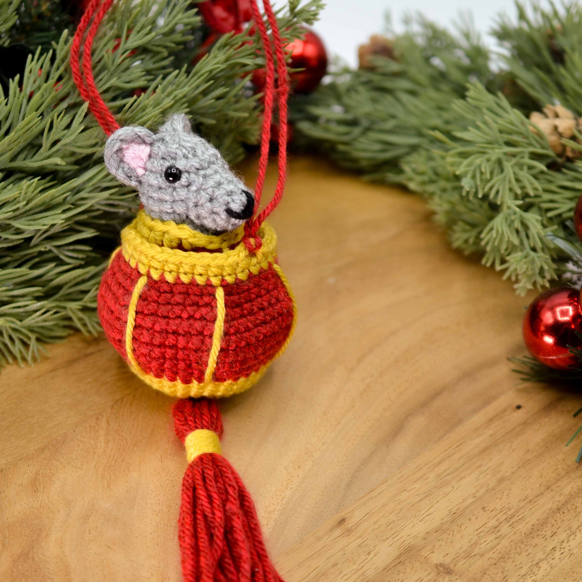 A crocheted mouse in a crocheted red lantern, placed on a wooden surface. The set is a lunar new year ornament. 