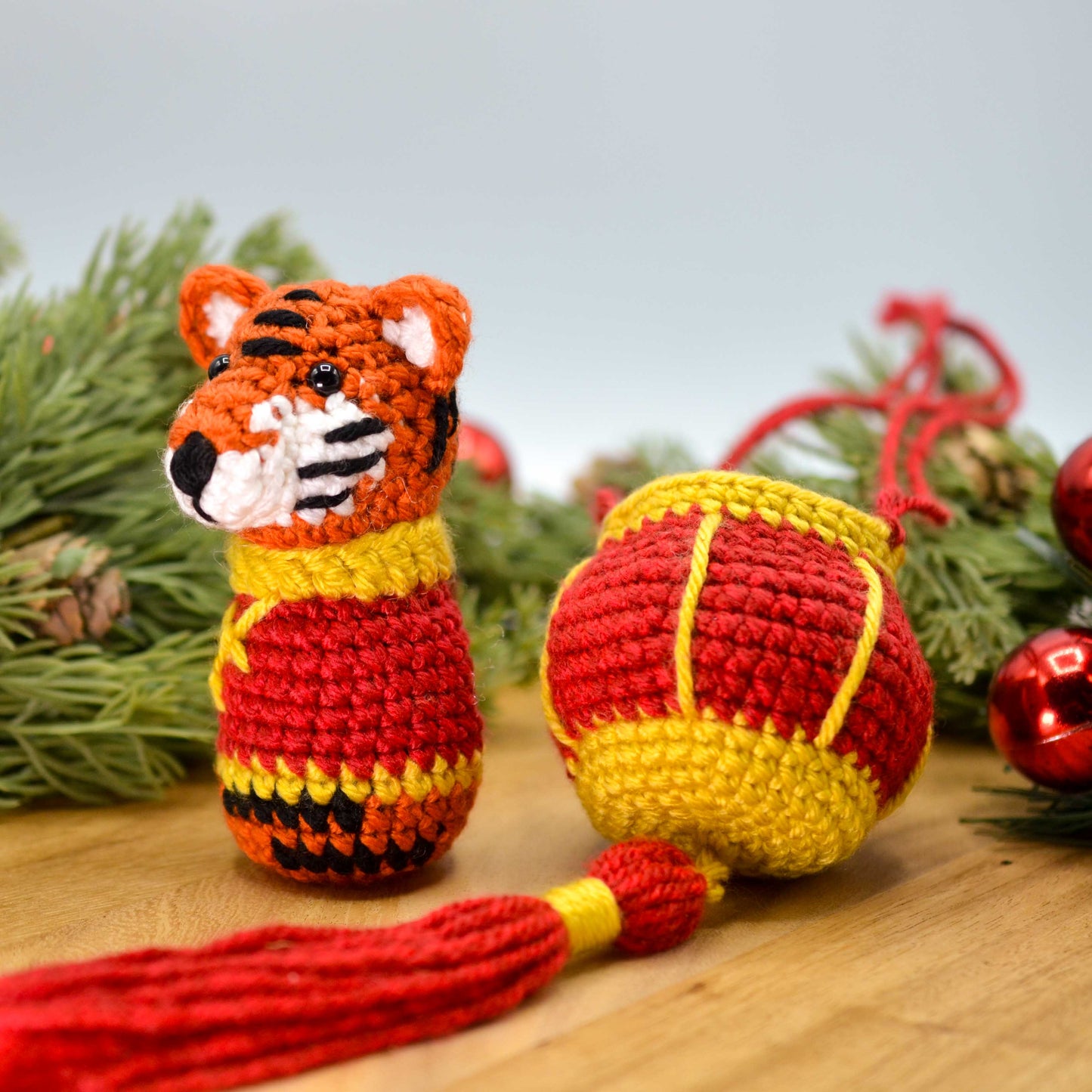 Crocheted tiger, facing front, and a red lantern on a wooden surface with festive decorations in the background.