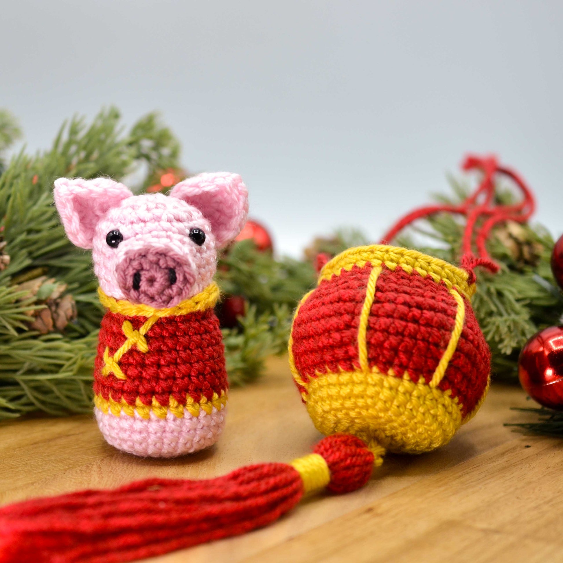 Crocheted pig ornament and red lantern displayed on a wooden surface. The pig is wearing a festive costume and facing front. 