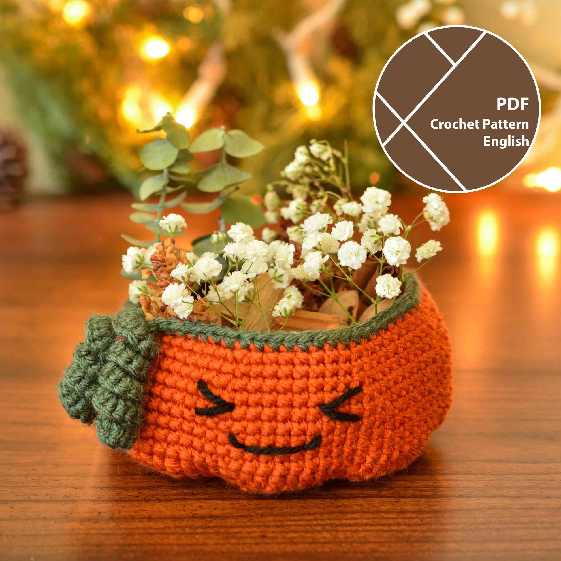 A crocheted pumpkin-shaped basket with a green stem and a smiling face. The basket is filled with white flowers and green leaves. The basket is sitting on a wooden surface. 