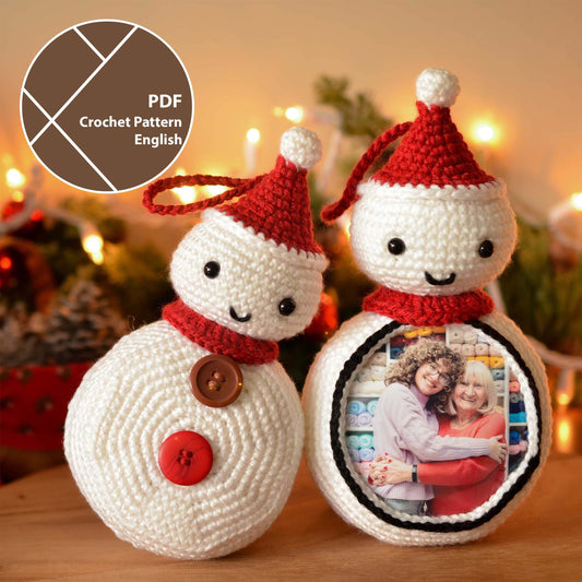 Two crocheted snowman ornaments with red Santa hats. One snowman has a photo frame on its front and the other snowman has two buttons on its front. The ornaments are on a wooden surface with a blurred background of Christmas lights.