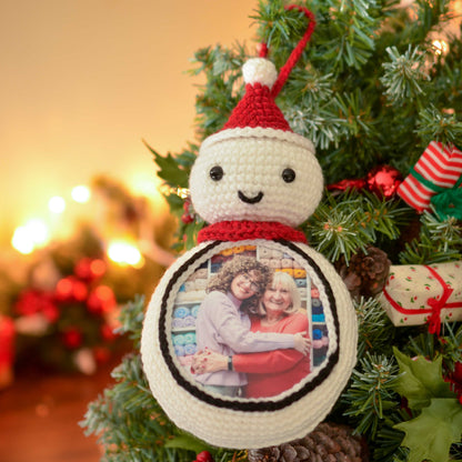 A white crocheted snowman ornament with a red Santa hat and a photo frame in the front. The photo frame contains a picture of two people smiling and hugging. The ornament is hanging on a green Christmas tree.