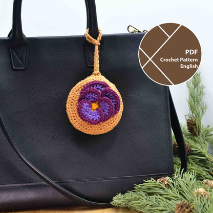 A crocheted pouch with a violet flower on the top. The pouch also doubles as a bag charm, hanging on a white black leather bag. 