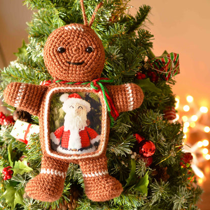 A crocheted gingerbread man ornament with a picture of Santa Claus inside a frame. The gingerbread man is brown with white icing details and is hanging on a green Christmas tree.
